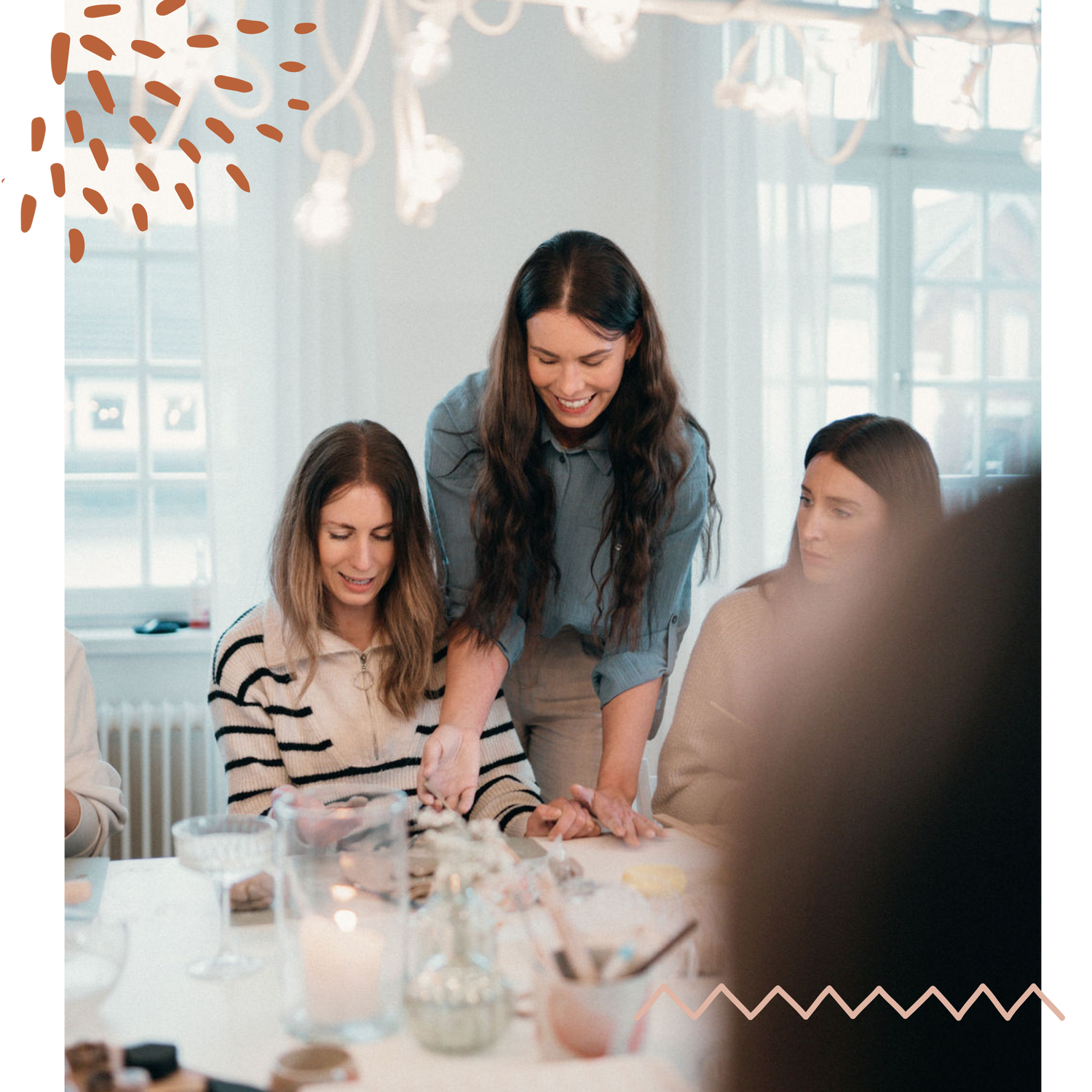 Eine Gruppe von Frauen nimmt an einem Töpfer-Workshop teil. Eine lächelnde Frau mit langen braunen Haaren steht unterstützend neben einer anderen Teilnehmerin und hilft ihr bei der Arbeit mit Ton. Der Raum ist hell und gemütlich, mit dekorativen Lichtern und verschiedenen Töpferutensilien auf dem Tisch. Die Szene strahlt eine freundliche und kreative Atmosphäre aus.