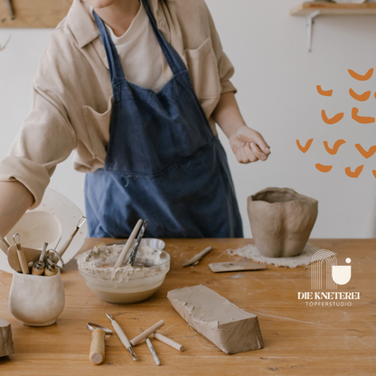 Person mit Schürze arbeitet an einem Holztisch mit Ton, Werkzeug und Keramik – Szene aus einem Töpferkurs.